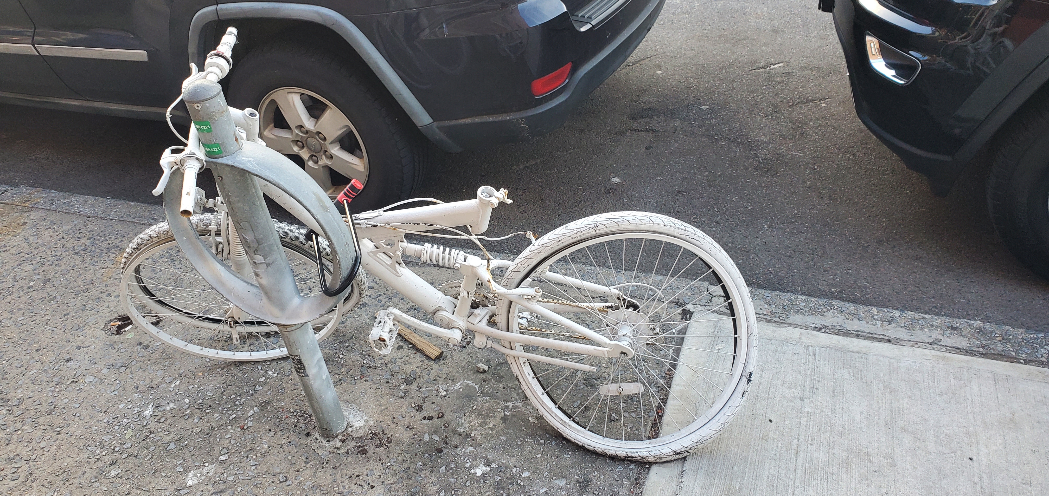 White bike with bent back tire chained to metal bike rack near parked cars