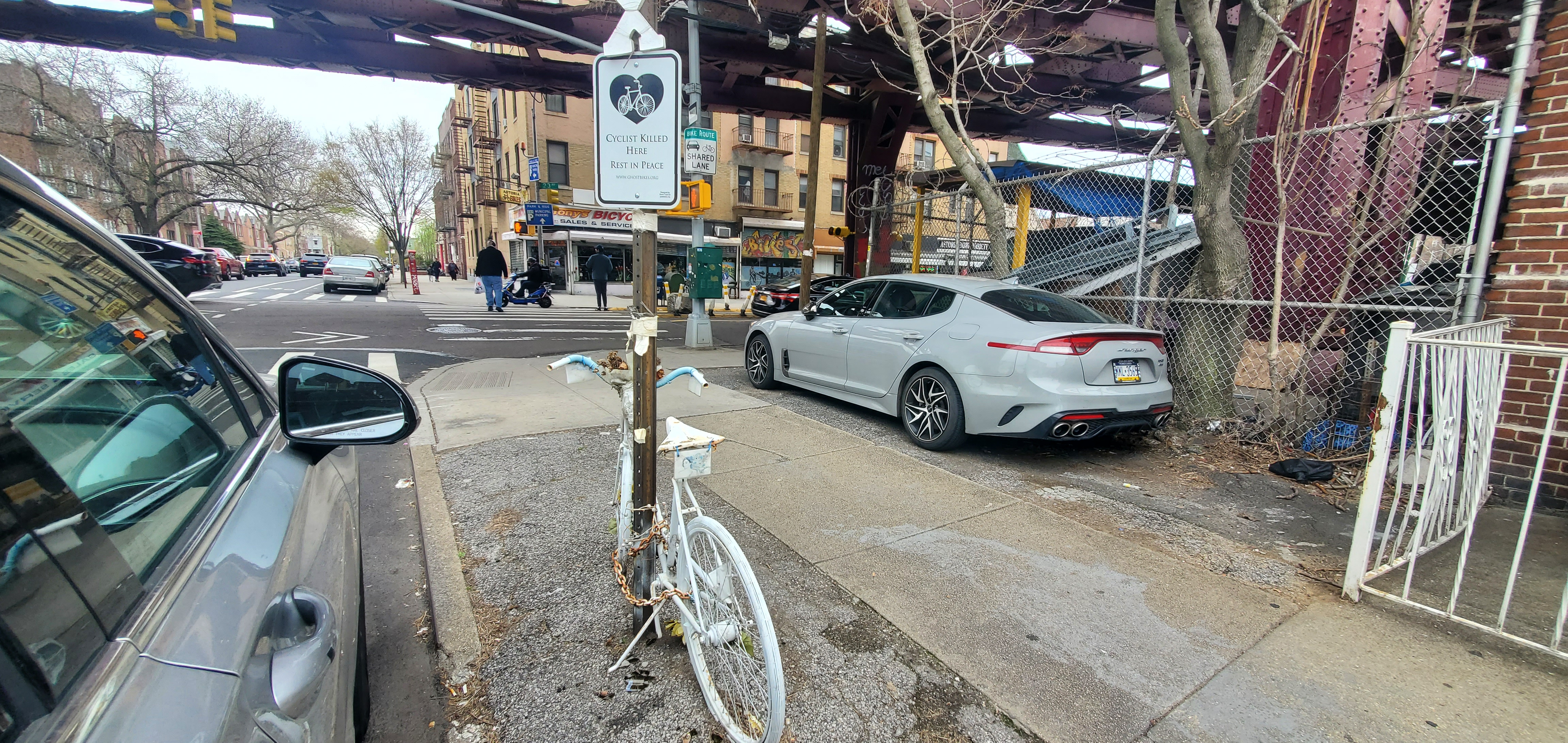 White bike and plaque chained to street sign between a car parked on the street and another parked on the sidewalk